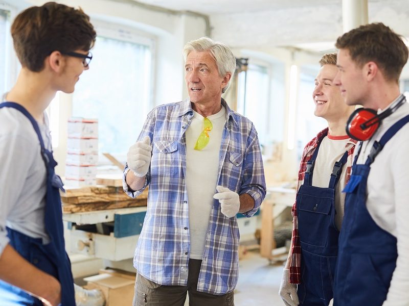 Confident handsome professional carpenter in work gloves standing in circle of young students and sharing skills with them, he showing carpentry workspace to interns