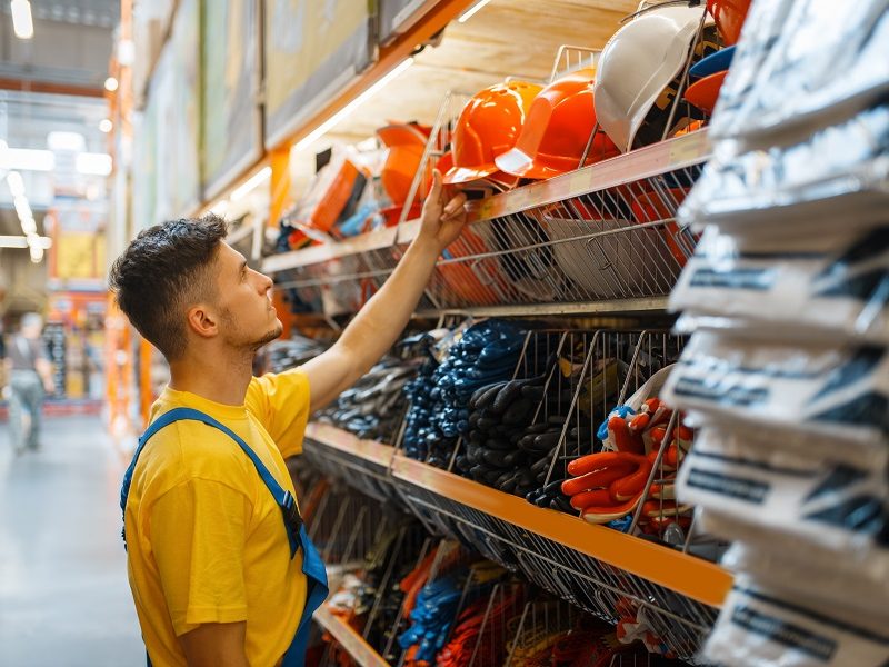 Male builder choosing helmet at the shelf in hardware store. Constructor in uniform look at the goods in diy shop