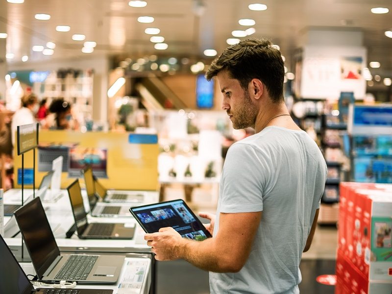 Side view of young male customer using tablet while searching for digital laptop in modern store with various gadgets