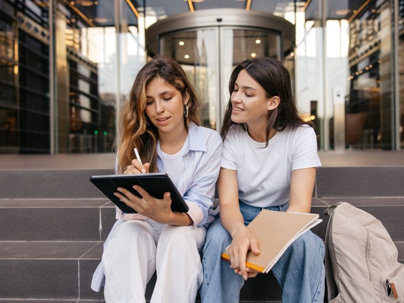 Calm young caucasian girls use tablet to search for information sitting on street. Blonde and brunette are preparing for lesson, wear casual clothes. Lifestyle concept